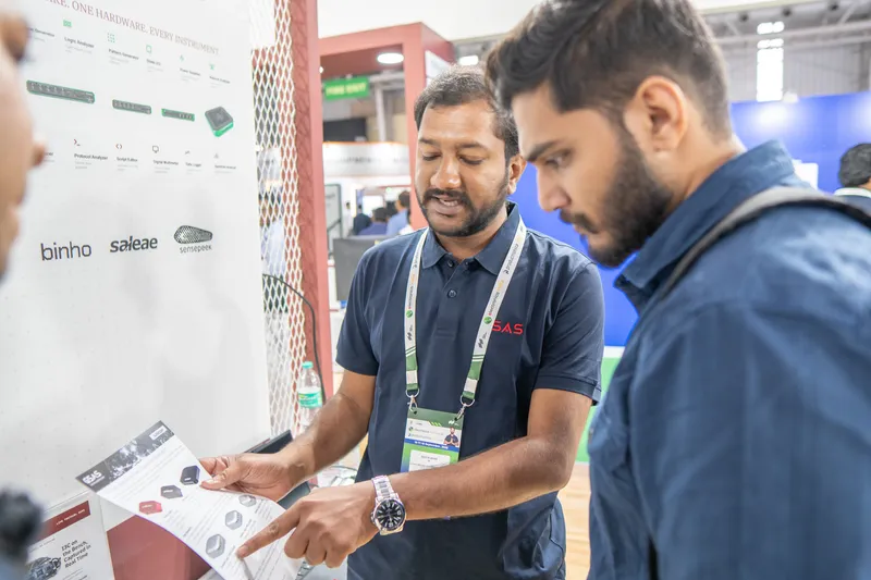 A GSAS engineer goes through a product leaflet with a visitor beside the Binho and Saleae wall, electronica India 2026